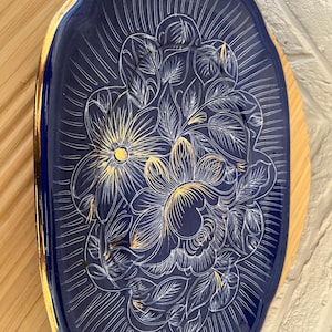 May include: A navy blue, oval decorative tray with a gold rim. The tray features a floral design in white and gold, with radiating lines. It rests on a light brown wooden surface. The tray's dimensions are not visible.