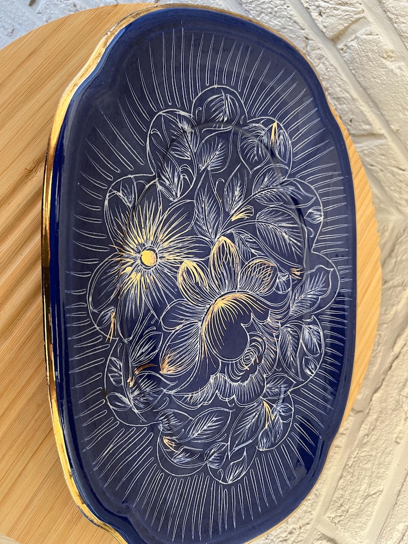 May include: A navy blue, oval decorative tray with a gold rim. The tray features a floral design in white and gold, with radiating lines. It rests on a light brown wooden surface. The tray's dimensions are not visible.