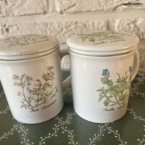 Può includere: Due tazze da tè in ceramica bianca con coperchi, con illustrazioni botaniche e testo. Una tazza mostra camomilla, l'altra menta. Le tazze hanno manici e sono su un vassoio verde.