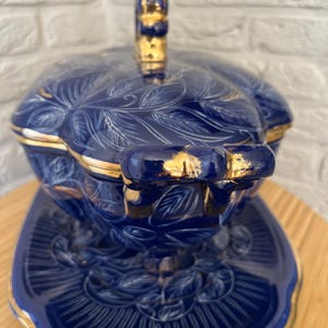 May include: A blue ceramic serving dish with a lid and a matching plate. The dish features a floral design with gold trim. The handle on the lid is also gold. The dish is sitting on a wooden surface.
