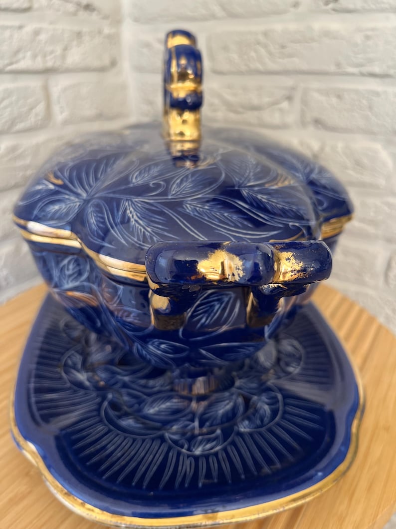 May include: A blue ceramic serving dish with a lid and a matching plate. The dish features a floral design with gold trim. The handle on the lid is also gold. The dish is sitting on a wooden surface.