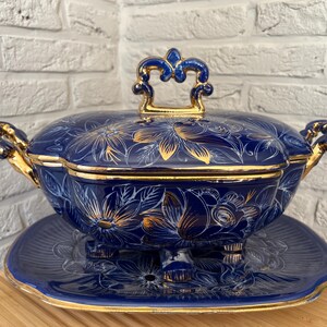 May include: A navy blue ceramic serving dish with a matching lid and a decorative plate. The dish and lid are decorated with gold floral patterns and trim. The handles and lid handle are gold.