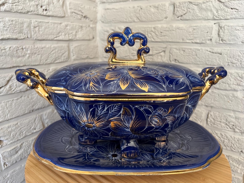 May include: A navy blue ceramic serving dish with a matching lid and a decorative plate. The dish and lid are decorated with gold floral patterns and trim. The handles and lid handle are gold.