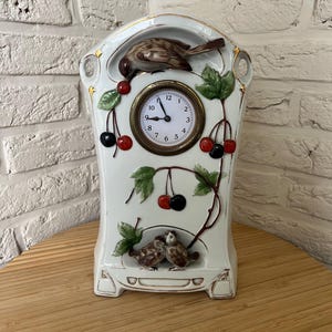 May include: A white ceramic mantel clock featuring a round analog face with black numerals and hands. The clock is decorated with three-dimensional brown birds, green leaves, and red and black cherries. Gold trim accents the edges of the clock.