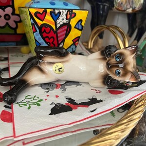 Puede incluir: Una figura de cerámica de un gato siamés con ojos azules, tumbado boca arriba. El gato tiene un cuerpo color crema con marcas marrones oscuras y descansa sobre una servilleta estampada. Otros objetos decorativos están en el fondo.