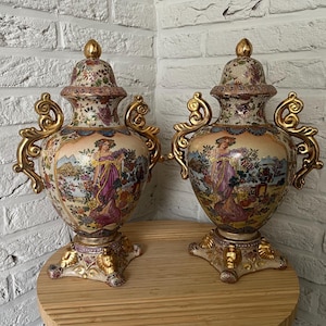 Może przedstawiać: Para pasujących ceramicznych urn z pokrywkami, wyposażonych w złote uchwyty w kształcie zwojów i ozdobne podstawy. Korpus każdej urny przedstawia kolorową ilustrację postaci w fioletowej sukni na tle krajobrazu, ozdobioną motywami kwiatowymi.