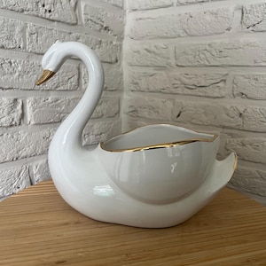 Puede incluir: Un cuenco de cerámica blanca con forma de cisne que presenta un acabado liso y brillante, con un borde pintado en oro a lo largo del pico y la parte superior del cuerpo. El cisne tiene un cuello largo y curvado y un centro hueco.