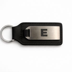 Puede incluir: Llavero de cuero negro con una placa de metal plateado. La placa presenta la letra "E" en negrita, sin serifa. El llavero tiene un llavero plateado y está sobre un fondo blanco.