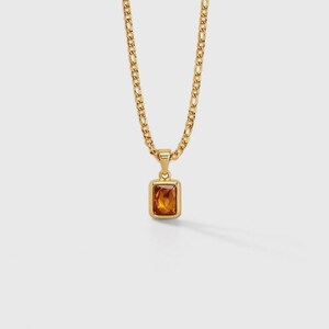 May include: A gold-colored necklace with a rectangular amber-colored pendant. The pendant is set in a gold frame and hangs from a gold chain. The chain has a curb link design. The necklace is displayed on a white background.