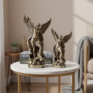 May include: Two bronze-colored statues of Saint Michael the Archangel, one large and one small, stand on a white and gold table. The statues depict a winged figure holding a sword and standing over a defeated figure. The statues are detailed and ornate.