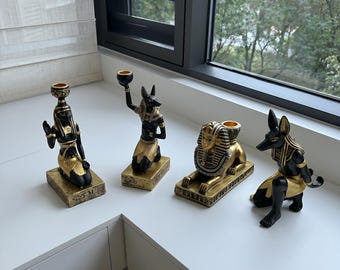 May include: Four Egyptian-themed candle holders in black and gold. The set includes figures of Anubis, a pharaoh, and other ancient Egyptian deities. Each holder has a small cup for a candle.