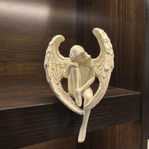 May include: A cream-colored angel figurine with detailed wings, seated with its head resting on its knees. The sculpture is placed on a dark brown wooden shelf, creating a contrast in color and texture. The angel's pose suggests contemplation.