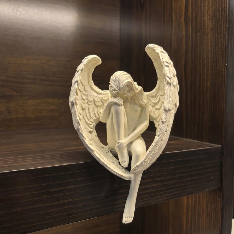 May include: A cream-colored angel figurine with detailed wings, seated with its head resting on its knees. The sculpture is placed on a dark brown wooden shelf, creating a contrast in color and texture. The angel's pose suggests contemplation.