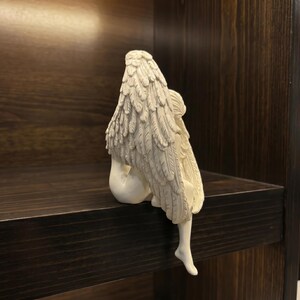 May include: A cream-colored angel figurine with detailed feather wings, seated on a dark wooden shelf. The angel is sculpted with delicate features and is positioned with its back to the viewer, creating a serene and contemplative pose. The wings are the most prominent feature.