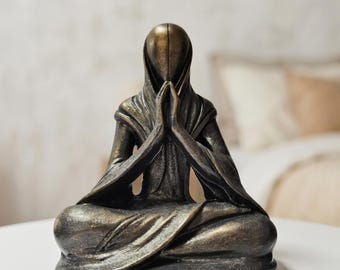May include: Bronze-colored statue of a figure in a meditative pose. The figure is seated with legs crossed, hands clasped in prayer, and a hooded garment. The statue has a smooth, metallic finish and is placed on a white surface.