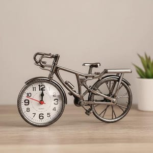 Puede incluir: Un reloj plateado con forma de bicicleta y esfera blanca. La esfera del reloj tiene números negros y manecillas rojas. La bicicleta tiene detalles, como manillar, asiento y ruedas. El reloj es un objeto decorativo.