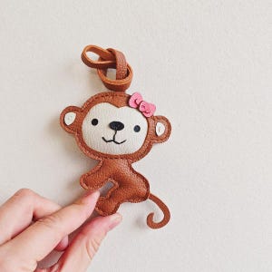 May include: A brown and cream-colored monkey-shaped keychain made of textured material. The monkey features a small pink bow on its head, black embroidered eyes, a black nose, and a curved tail, attached to a brown loop for hanging.
