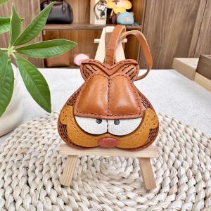 May include: A leather coin purse in the shape of a cartoon cat's face. The purse is made of brown, tan, white, and pink leather, with a small handle. It is displayed on a small wooden stand.