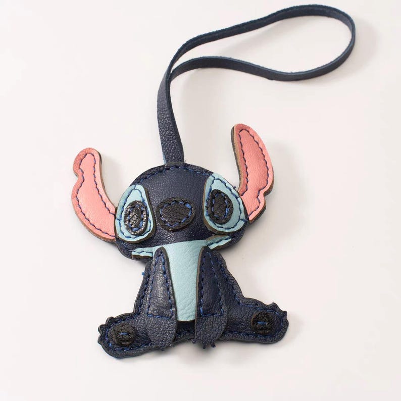 May include: A navy blue leather Stitch keychain. The keychain features Stitch's face with light blue eyes, pink ears, and a light blue belly. The keychain has a navy blue loop for attaching to keys or a bag.
