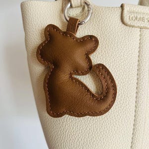 May include: A brown leather cat-shaped keychain charm with stitched detailing, hanging from a silver ring attached to a cream-colored leather handbag. The bag has a textured surface and a top handle.