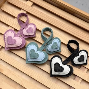 May include: Three heart-shaped keychains made of textured leather. Each keychain features a layered design with a smaller heart stitched onto a larger heart, attached to a circular loop. The colours are purple and pink, teal and light blue, and black and white.