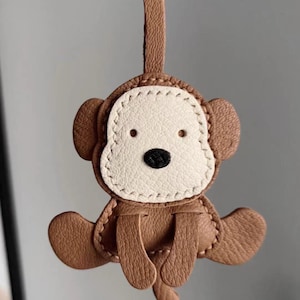 Op de afbeelding: Een bruine leren aap sleutelhanger met een witte gezicht en zwarte neus. De sleutelhanger heeft een lus aan de bovenkant voor bevestiging en is gestikt met witte draad. De aap zit met uitgestrekte benen.