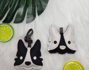 May include: Two leather keychains shaped like a dog's face, one with a loop and strap. The keychains are black and white with stitched details. The background is a soft, white surface with a green leaf and a lime slice.