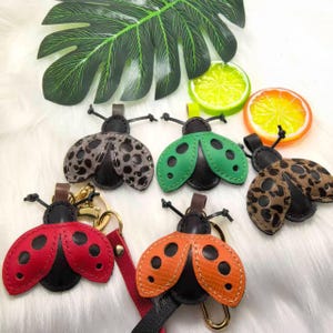 May include: A collection of ladybug-shaped keychains in various colors, including red, orange, green, and leopard print. Each keychain features a black body and a small loop for attaching keys. The keychains are displayed on a white, fluffy surface.