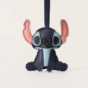 May include: A navy blue leather Stitch character keychain. The keychain features Stitch's signature large ears, blue eyes, and light blue accents. The keychain is hanging from a navy blue leather strap.
