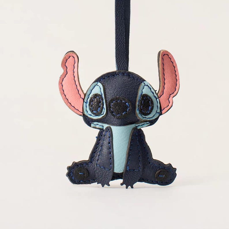 May include: A navy blue leather Stitch character keychain. The keychain features Stitch's signature large ears, blue eyes, and light blue accents. The keychain is hanging from a navy blue leather strap.