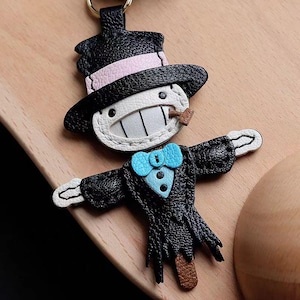 May include: A black leather keychain featuring a cartoon character with a top hat, a light pink band, and a blue bow tie. The character has a white face, a wide grin, and a small brown object in its mouth. The keychain is on a wooden surface.