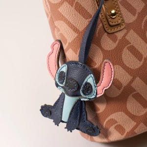May include: A blue and pink Stitch character charm attached to a brown tote bag. The charm is made of leather and features Stitch's signature large ears and eyes. The tote bag has a repeating pattern of the letter "D".
