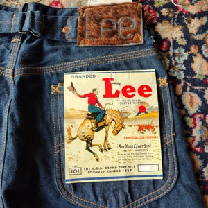 Può includere: Jeans in denim blu scuro con una toppa in finta pelliccia marrone con il logo Lee. Un'etichetta rettangolare in carta mostra un cowboy su un cavallo, il marchio Lee e testo che indica denim da 11,5 once e rivetti in rame.