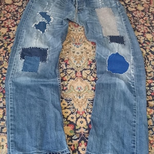 May include: Blue denim jeans featuring a distressed finish with multiple patches in varying shades of blue and grey. The patches are secured with visible white stitching, and the leg hems show frayed edges and wear throughout the fabric.