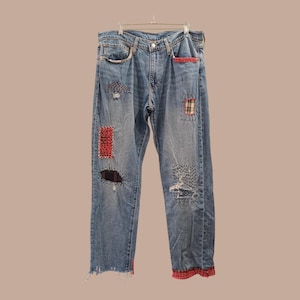 Può includere: Jeans di denim blu chiaro con diverse toppe rosse, nere e a quadri. I jeans presentano dettagli usurati e bordi sfilacciati. I jeans sono appesi a una gruccia di metallo.
