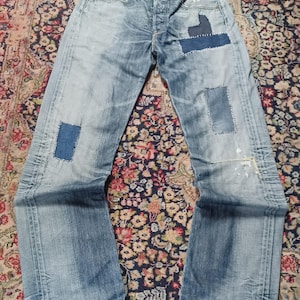 Può includere: Jeans in denim azzurro con chiusura a bottoni, diversi inserti rettangolari in tessuto di varie tonalità di blu, cuciture bianche visibili attorno alle toppe e lievi schizzi di vernice sulla parte inferiore della gamba sinistra.