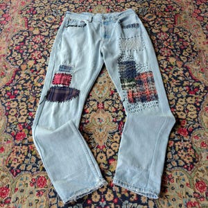 Op de afbeelding: Lichtblauwe spijkerbroek met diverse rechthoekige stoffen patches in ruitpatronen en effen kleuren. De patches zijn bevestigd met zichtbare donkere stiksels, wat zorgt voor een textuurrijk en hersteld uiterlijk op de voorzijde van de broekspijpen.