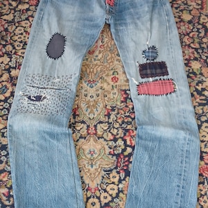 Può includere: Jeans in denim azzurro con chiusura a bottoni e una toppa cucita rosa. Le gambe sono decorate con varie toppe in tessuto blu, a quadri e rosa, insieme a dettagli ricamati a mano visibili e orli sfrangiati.