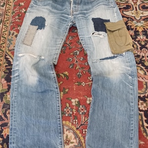 Könnte beinhalten: Hellblaue Jeans aus Denim mit Knopfleiste, abgenutzten Stellen und mehreren Flicken. Das rechte Bein hat einen dunkelblauen und grauen Flicken, während das linke Bein einen dunkelblauen Flicken und eine beige Cargotasche mit Klappenverschluss aufweist.