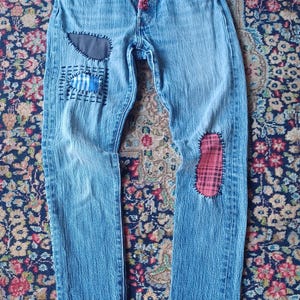Könnte beinhalten: Hellblaue Jeans aus Denim mit Knopfleiste und rotem Stoffakzent, einem dunkelgrauen Flicken auf dem rechten Oberschenkel, einem blau-weiß gestreiften Flicken darunter und einem rot karierten Flicken auf dem linken Knie, befestigt mit schwarzen Nähten.