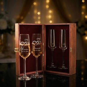 May include: A wooden box with two champagne flutes filled with a golden liquid. The flutes are engraved with a heart design, names, and a date. Two additional flutes are inside the box. The box has a red velvet interior.