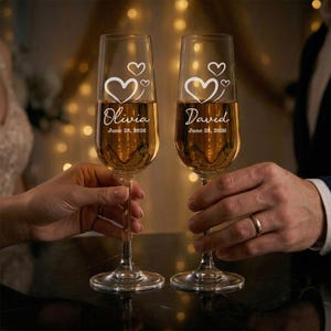 Könnte beinhalten: Zwei Champagnergläser gefüllt mit einer goldenen Flüssigkeit. Jedes Glas ist mit einem Herzmotiv und einem Namen, "Olivia" und "David", sowie dem Datum "28. Juni 2026" graviert. Die Gläser werden von zwei Personen gehalten.