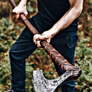 May include: Axe with a wooden handle and a metal blade. The handle is wrapped with leather straps. The axe is held by a person wearing a black shirt and dark blue pants.