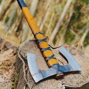 Op de afbeelding: Een dubbelzijdige bijl met een houten handvat en een zwarte leren wikkel. De bijlkoppen zijn zilverkleurig en het handvat is lichtbruin. De bijl ligt op een boomstronk.