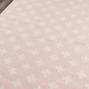 May include: A pink rug featuring a repeating pattern of white crosses. The rug has a soft texture and a simple, modern design. Suitable for a living room or bedroom, the rug is made of a soft material.