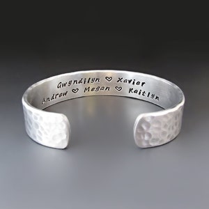 May include: A silver-toned cuff bracelet with a hammered texture on the exterior. The interior is engraved with the names Gwyndilyn, Xavier, Andrew, Megan, and Kaitlyn, separated by small heart symbols in a clear, dark, sans-serif font.