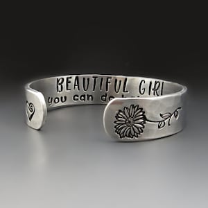 May include: Silver cuff bracelet with the words "Beautiful Girl You Can Do Anything" engraved on it. A small heart and a flower are also engraved on the bracelet.
