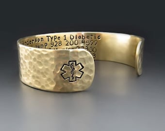 Puede incluir: Un brazalete de metal dorado con textura martillada y un símbolo médico negro. El interior está grabado con el texto: Anderson Type 1 Diabetic, pump 928 200 4577 y números adicionales. El brazalete tiene un diseño abierto en los extremos.