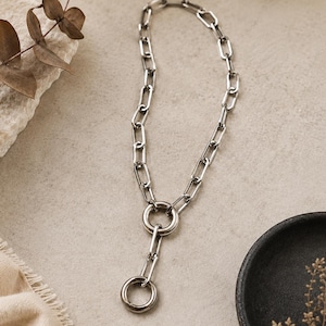 May include: A silver-toned necklace featuring elongated rectangular chain links. The design includes a central circular ring connector that joins the main chain to a shorter vertical segment, which terminates in a smaller circular ring pendant.