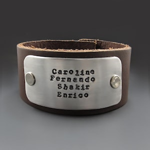 May include: Brown leather cuff bracelet with a silver metal plate engraved with the names Caroline, Fernando, Shakir, and Enrico.